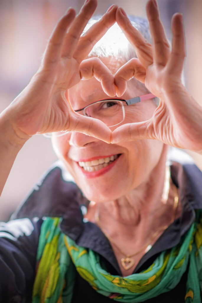 A smiling senior woman with glasses and a green scarf making a heart gesture with her hands, symbolizing joy and emotional support during hospice care.
